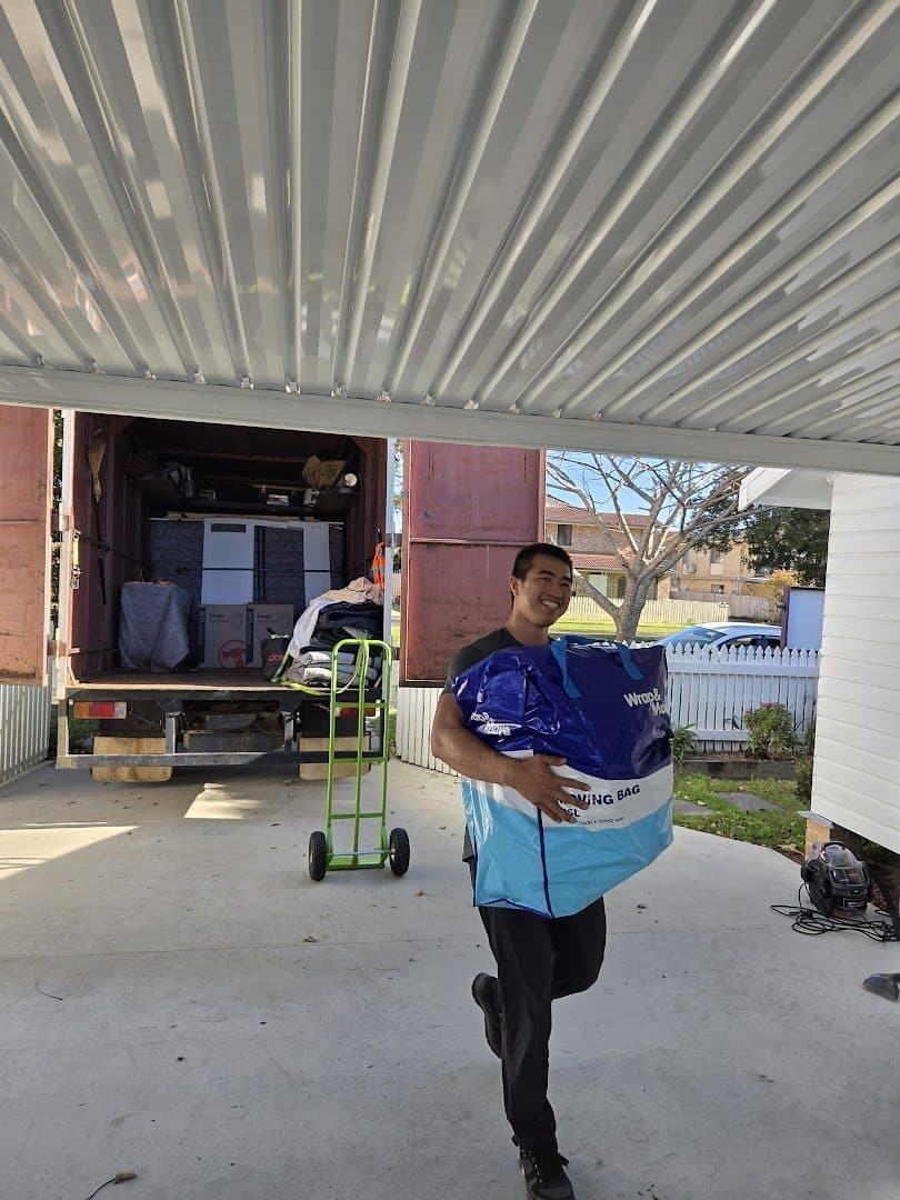 Crew member carrying a large moving bag under a carport, loaded truck behind