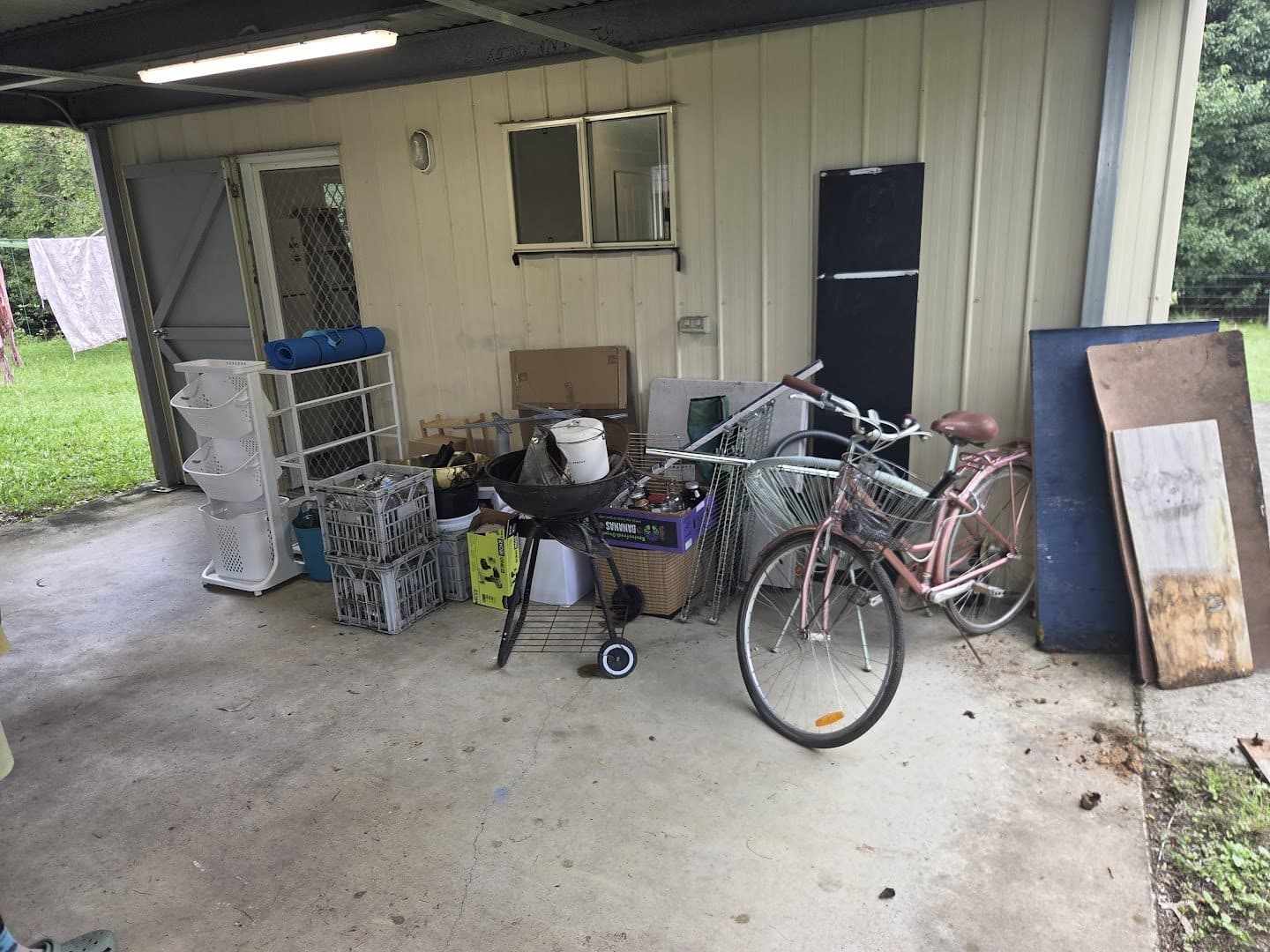 A carport staged with clear-out items: bikes, a kettle barbecue, crates and boards